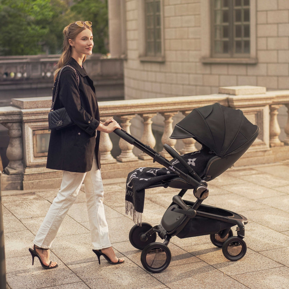 Mima Creo Stroller in black with expansive canopy being pushed along a city promenade by a stylish woman.