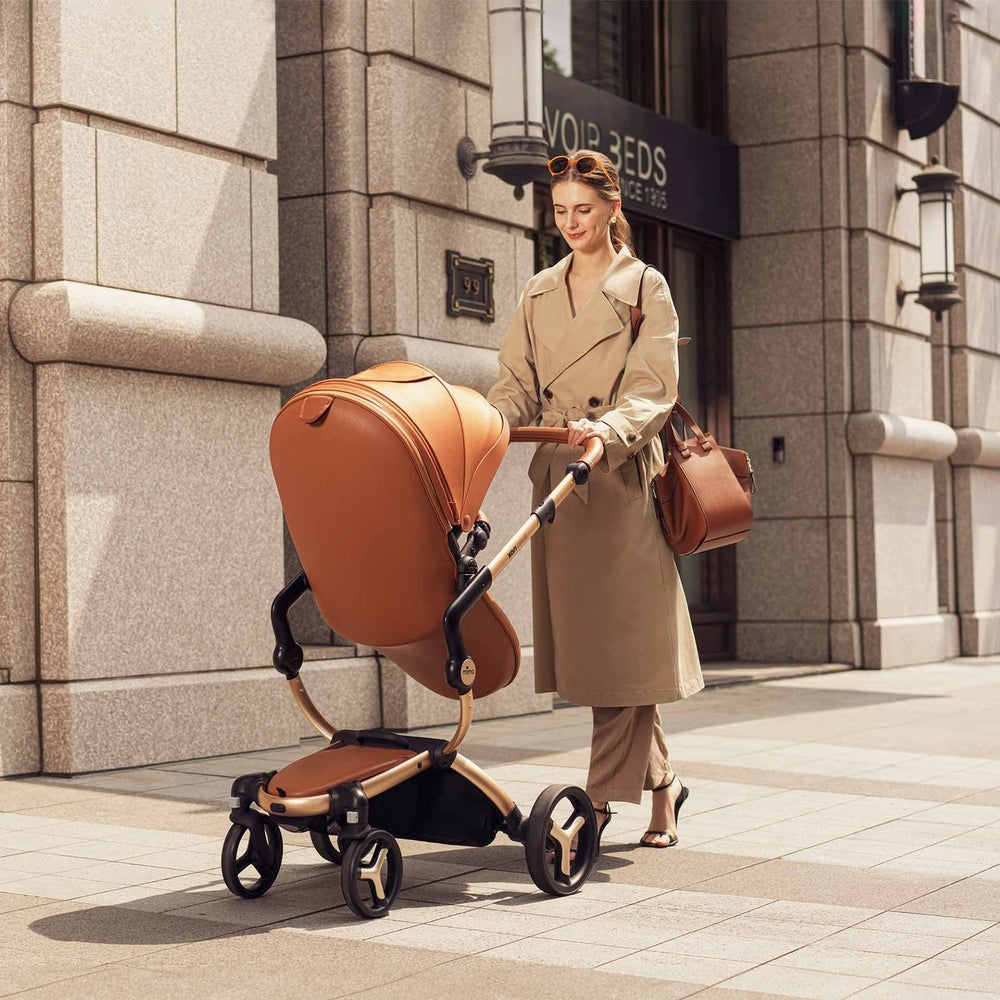Mima Xari Max stroller with tan bassinet, a stylish woman pushes the baby carriage along a city sidewalk.