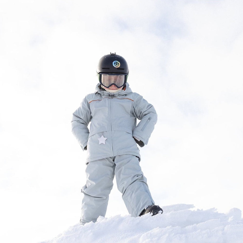 MINI A TURE Matadane Ski Tech Jacket Grs Quarry worn by a child standing on snowy ground in a light gray ski suit with helmet and goggles