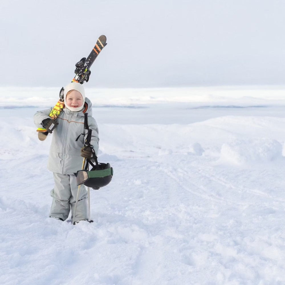 MINI A TURE Matadane Ski Tech Jacket Grs Quarry worn by a smiling child in a snowy landscape, carrying skis and helmet.