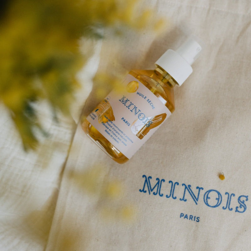Minois Paris dry oil bottle, 150 ml, amber glass with white spray cap on beige cloth bearing blue MINOIS PARIS logo