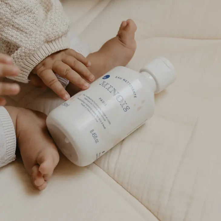 Minois Paris Cleansing Water 250 ml bottle beside a baby's feet on soft beige bedding