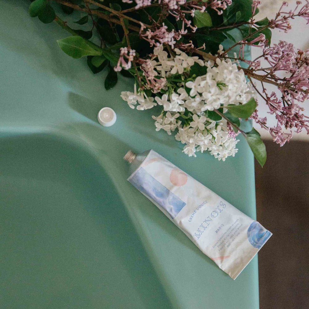 Minois Paris Face and Body Cream 100 ml tube on green surface beside white and pink lilac flowers