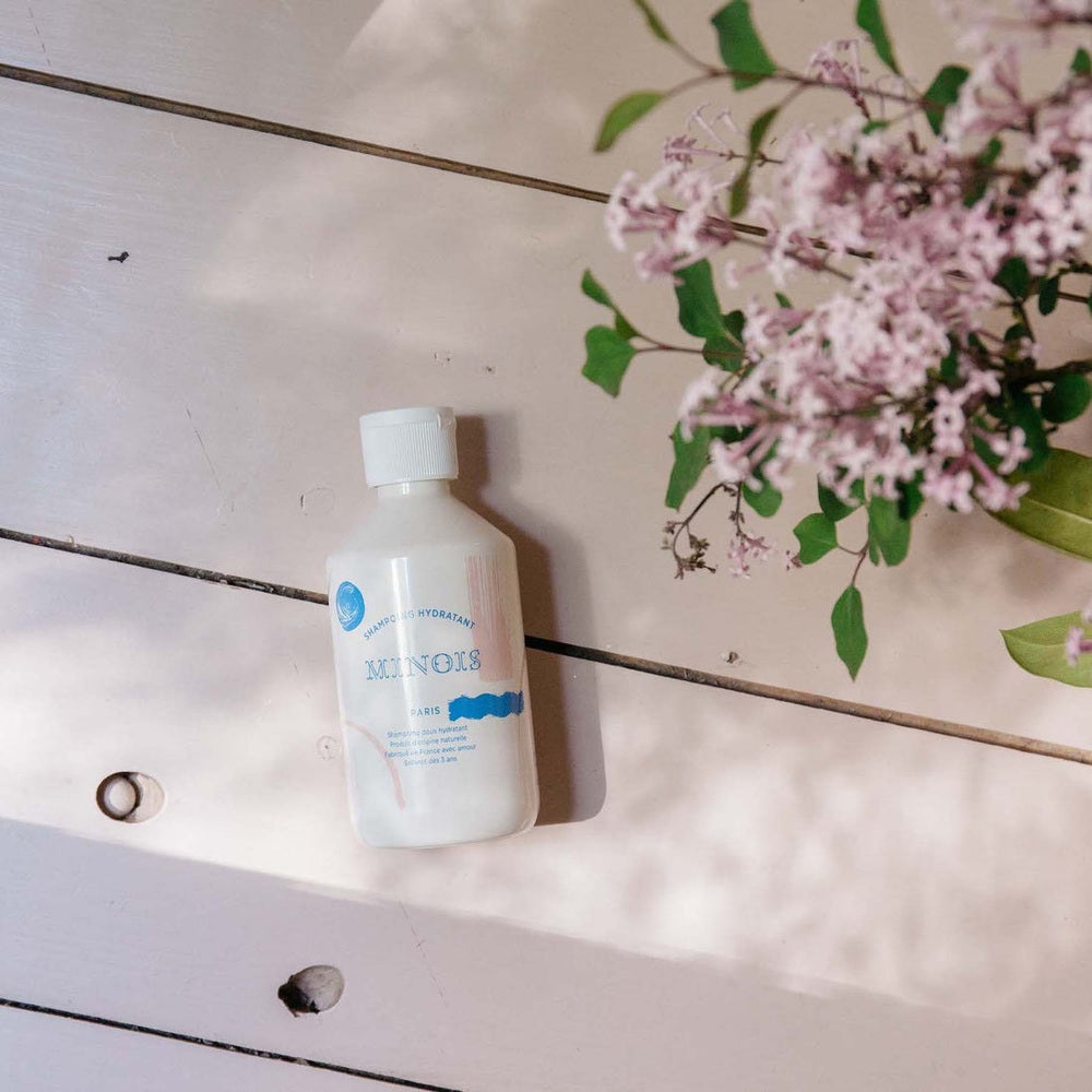 Minois Paris hydrating shampoo bottle 300 ml, white plastic on pale wooden table beside pink blossoms