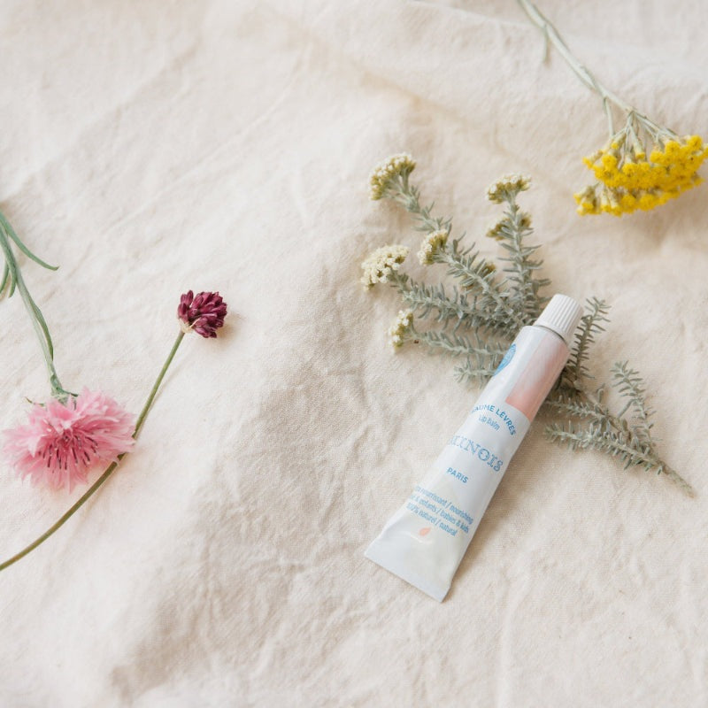 Minois Paris Lip Balm 12 ml in pink tube with white cap, displayed on beige fabric with flowers.