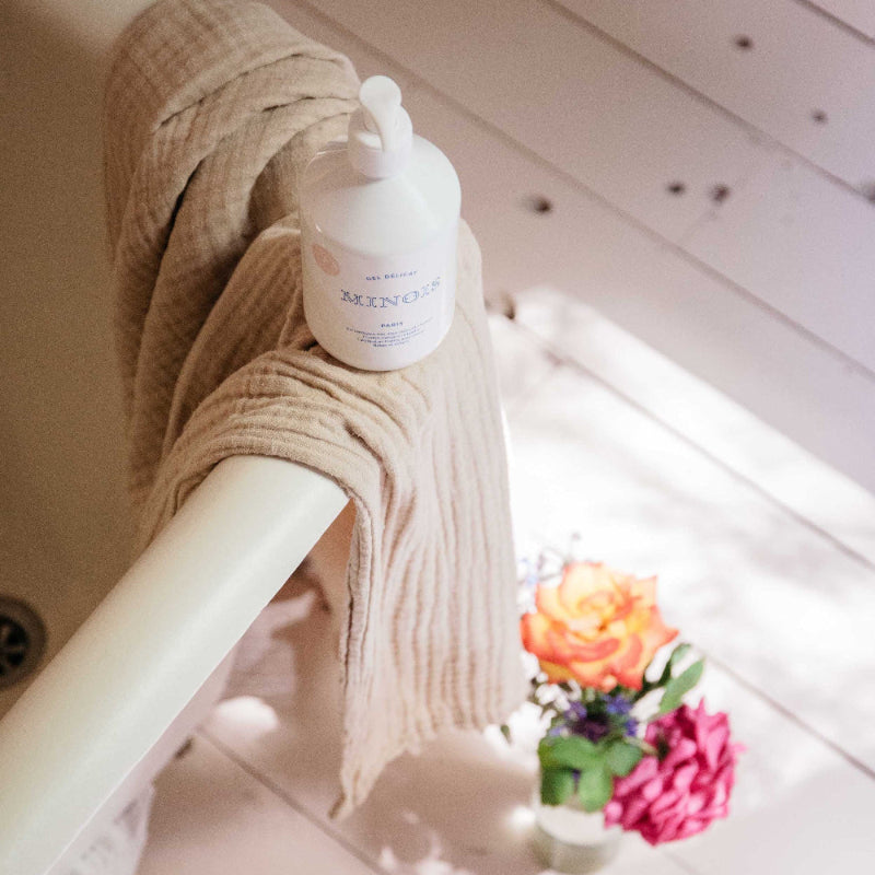 Minois Paris washing gel 500 ml on a beige knit blanket beside a white railing, with bouquet in background.