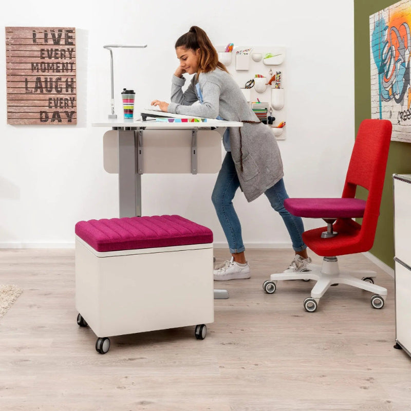 MOLL Champion Compact Express Height Adjustable Desk in a bright office; pink ottoman on wheels and red rolling chair, user leaning over desk.