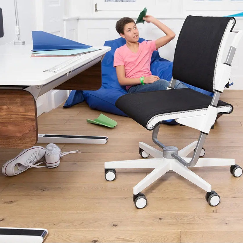 Moll Scooter Children Swivel Chair, white frame with black mesh seat, rolling on caster wheels beside a desk.