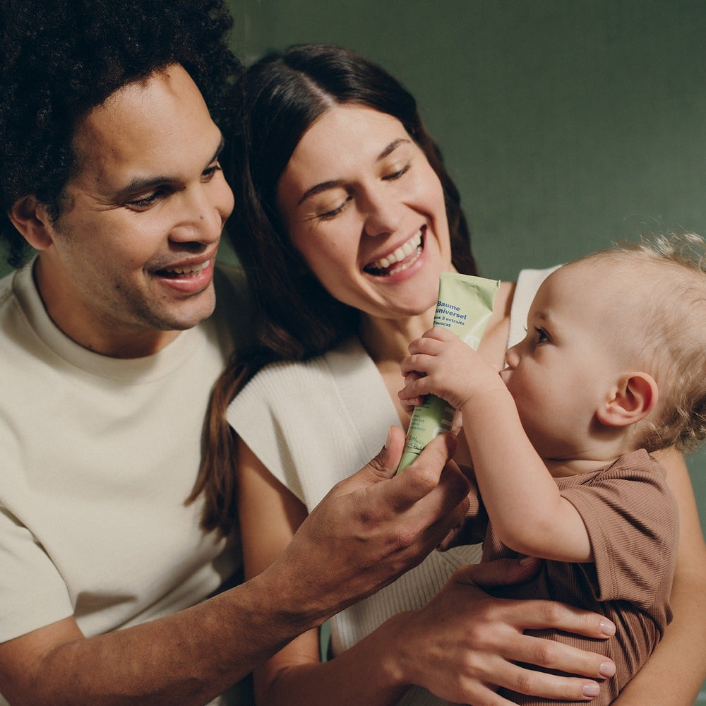 Mustela Organic Universal Balm With 3 Avocado Extracts 75 ml; parents smile as baby grasps the avocado balm tube.