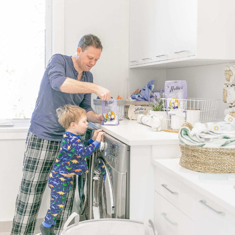 Two Nellie's Baby Laundry Soda tins on a white kitchen counter as a father and child load a washing machine