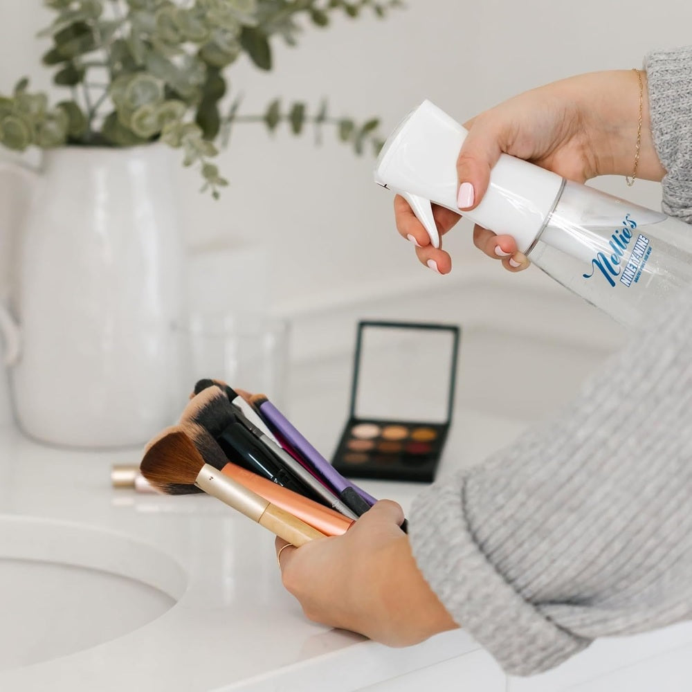 Nellie's Ninety-Nine spray bottle with blue logo beside makeup brushes on a white bathroom counter.