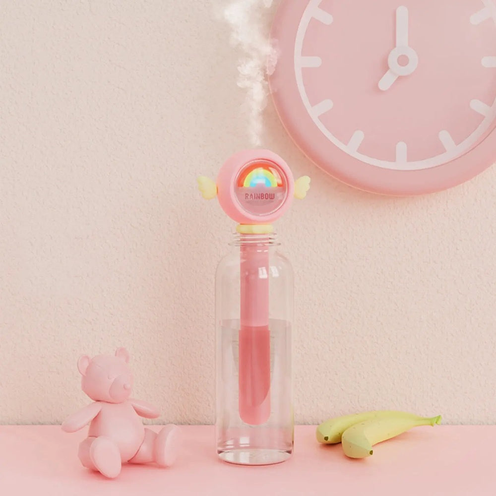 Nemibaby Rainbow Fairy Humidifier on a pink desk; clear glass bottle base, rainbow-cap top, tiny winged fairy.