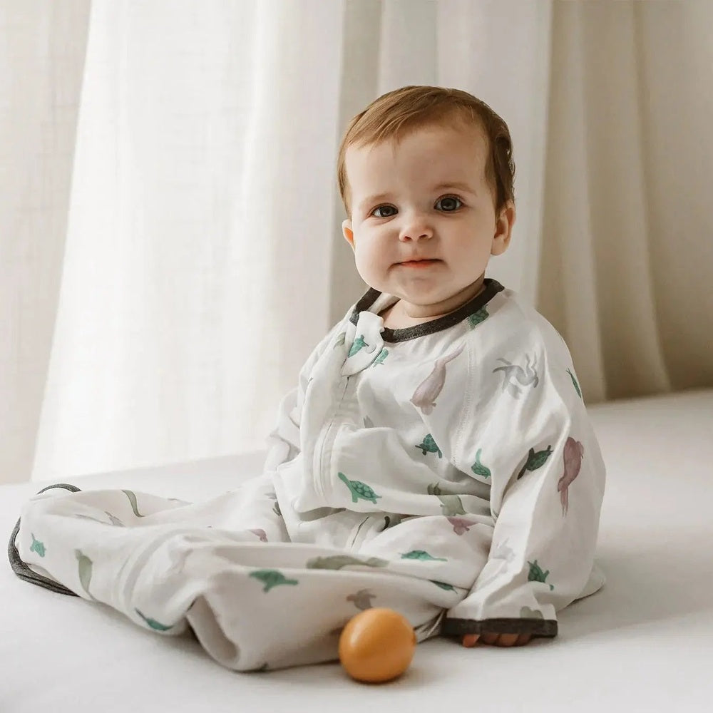 Nest Designs 0.23 Tog Bamboo Sleep Bag The Tortoise & The Hare; baby sits in a white 3/4-sleeve sleepsack with tortoise/hare print.