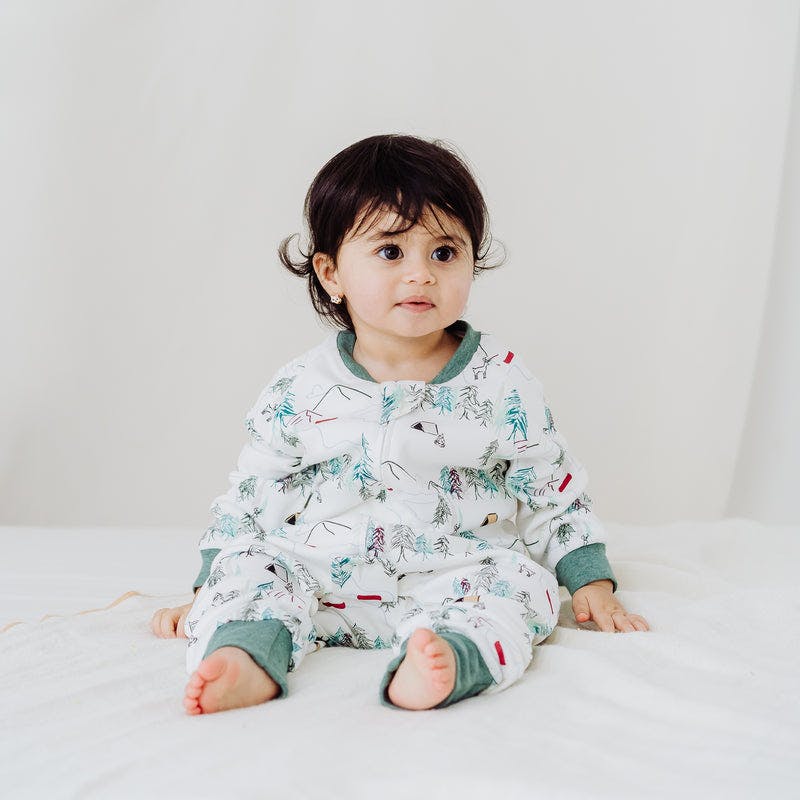 Nest Designs Avocado Bamboo Long Sleeve Footed Sleep Bag 1.0 Tog—baby sits on bed in forest-print sleep sack.