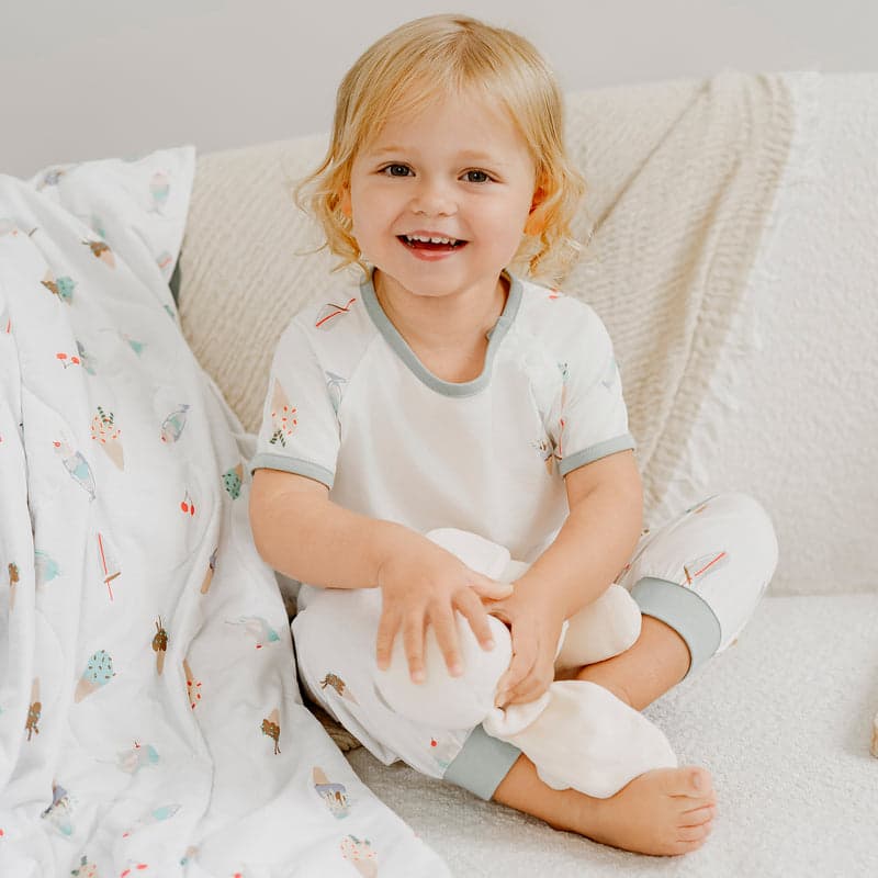 Nest Designs Avocado Bamboo Short Sleeve Play Set Ice Cream Summer; smiling toddler sits on couch in white pajamas with gray trim.