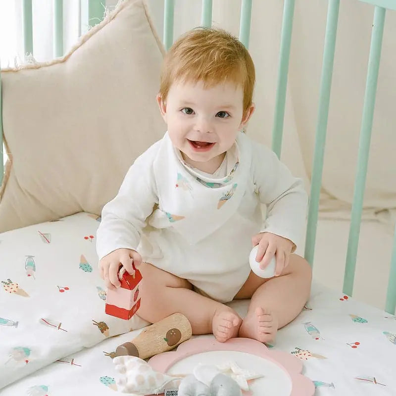 Nest Designs Bamboo Baby Bandana Bib One Size (3M-24M) in white with pastel print, worn by a smiling baby in a crib.