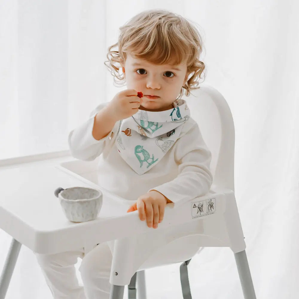 Nest Designs Bamboo Baby Bandana Bib One Size (3M-24M) in white with teal dinosaur print; toddler wears it while feeding.