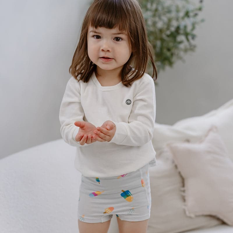 Nest Designs Bamboo Girls Underwear 2 Pack Sweet Treats; toddler stands indoors in white top and pastel shorts, cupping hands.