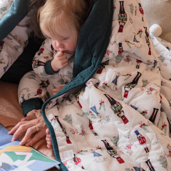 Nest Designs Bamboo Jersey Cozy Blanket Medium Nutcracker, teal-trimmed, kid snuggled as parent reads nearby.