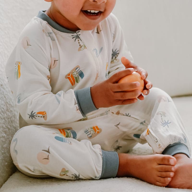 Nest Designs Bamboo Jersey One-Piece Zip Footless Sleeper Pastel Waves worn by a smiling baby, who is holding an orange toy.