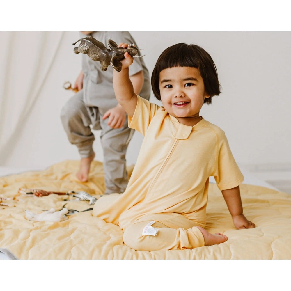 Nest Designs Bamboo Jersey Short Sleeve Footed Sleep Bag 0.5 Tog Pantone Sunset Gold, toddler sits on a bed waving a dinosaur.