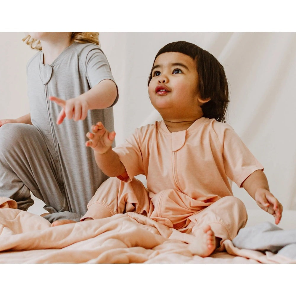 Nest Designs bamboo jersey short-sleeve footed sleep bag Pantone Bellini peach, toddler sits on blanket wearing it.