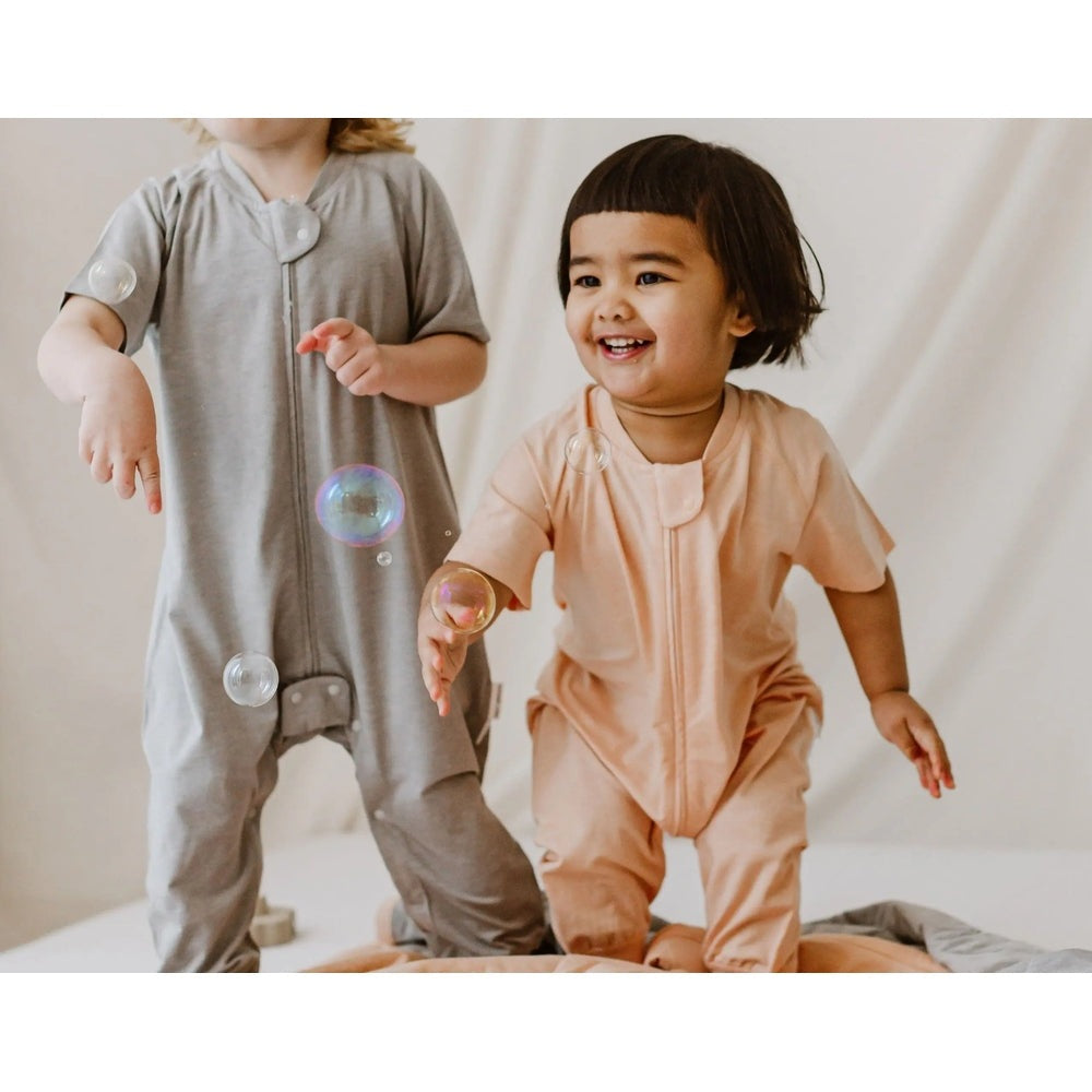 Nest Designs Bamboo Jersey Short Sleeve Footed Sleep Bag 0.5 Tog Pantone Bellini peach on a smiling baby reaching for bubbles.