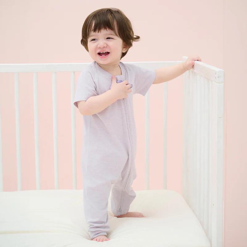 Nest Designs Bamboo Jersey Short Sleeve Slant Opening Romper Lavender Blue; toddler stands in a crib wearing the soft lavender one-piece.