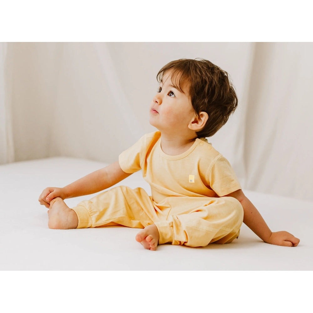 Nest Designs Bamboo Jersey Short Sleeve T-Shirt Pantone Sunset Gold: toddler sits cross-legged on white floor in soft yellow outfit.