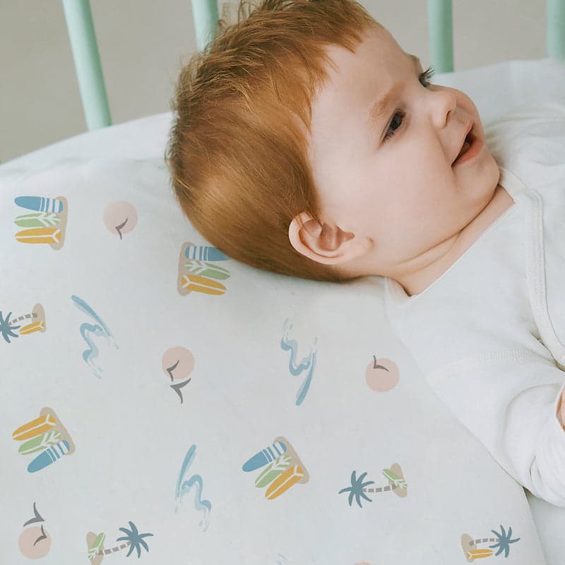 Nest Designs Bamboo Jersey Toddler Pillow with pastel waves pillowcase supports a smiling toddler on a white bed.