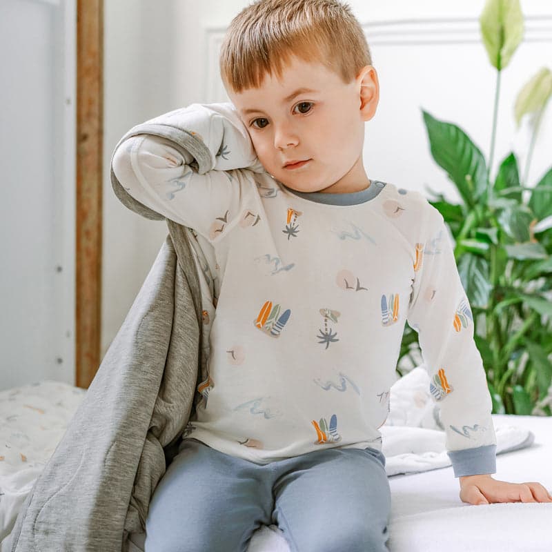 Nest Designs Bamboo Jersey Two-Piece Long Sleeve PJ Set Pastel Waves; child sits on bed wearing white top with pastel surf motifs and blue pants.