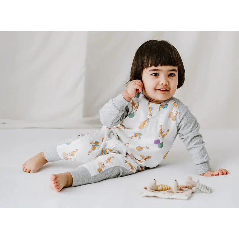 Nest Designs Bamboo Long Sleeve Footed Sleep Bag 1.0 Tog in giraffe print, baby sits calmly on white floor.