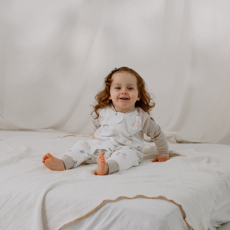Nest Designs Bamboo Long Sleeve Footed Sleep Bag 1.0 TOG; smiling baby sits upright on a white bed in the sleep bag.