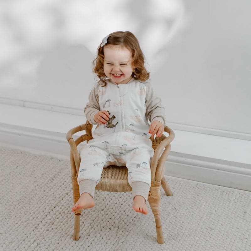 Nest Designs Bamboo Long Sleeve Footed Sleep Bag 1.0 Tog in cream with small print, toddler smiling while seated in a wicker chair.