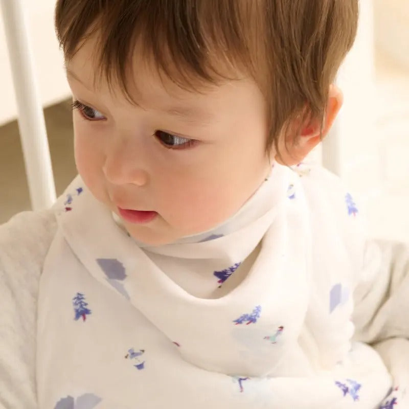 Nest Designs Bamboo Muslin Baby Bandana Bib Frosty Fun; white bib with blue trees, baby wearing it and looking aside.