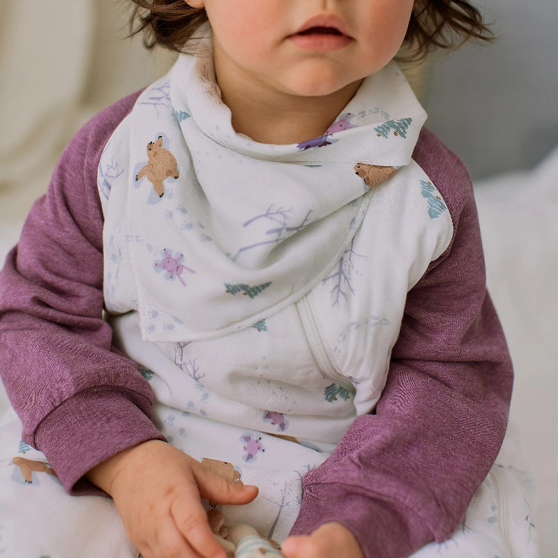 Nest Designs Bamboo Muslin Baby Bandana Bib Snow Angels, white with pastel prints; baby wears the bib around her neck.