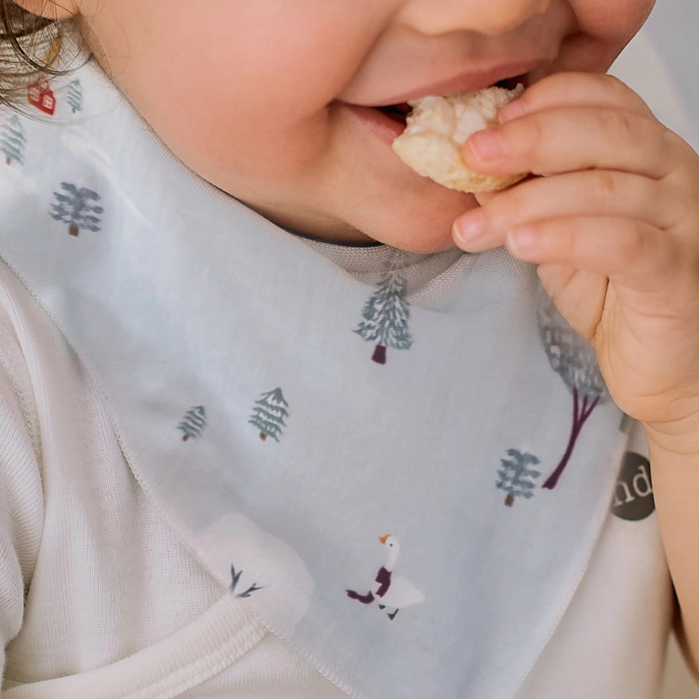Nest Designs Bamboo Muslin Baby Bandana Bib Snowy Snuggles bib worn by baby while munching a snack.