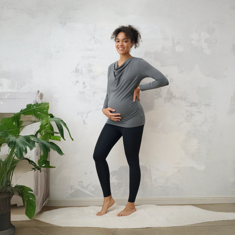 Pregnant woman in grey draped-neck top wearing black bamboo maternity leggings, standing indoors on a white rug.