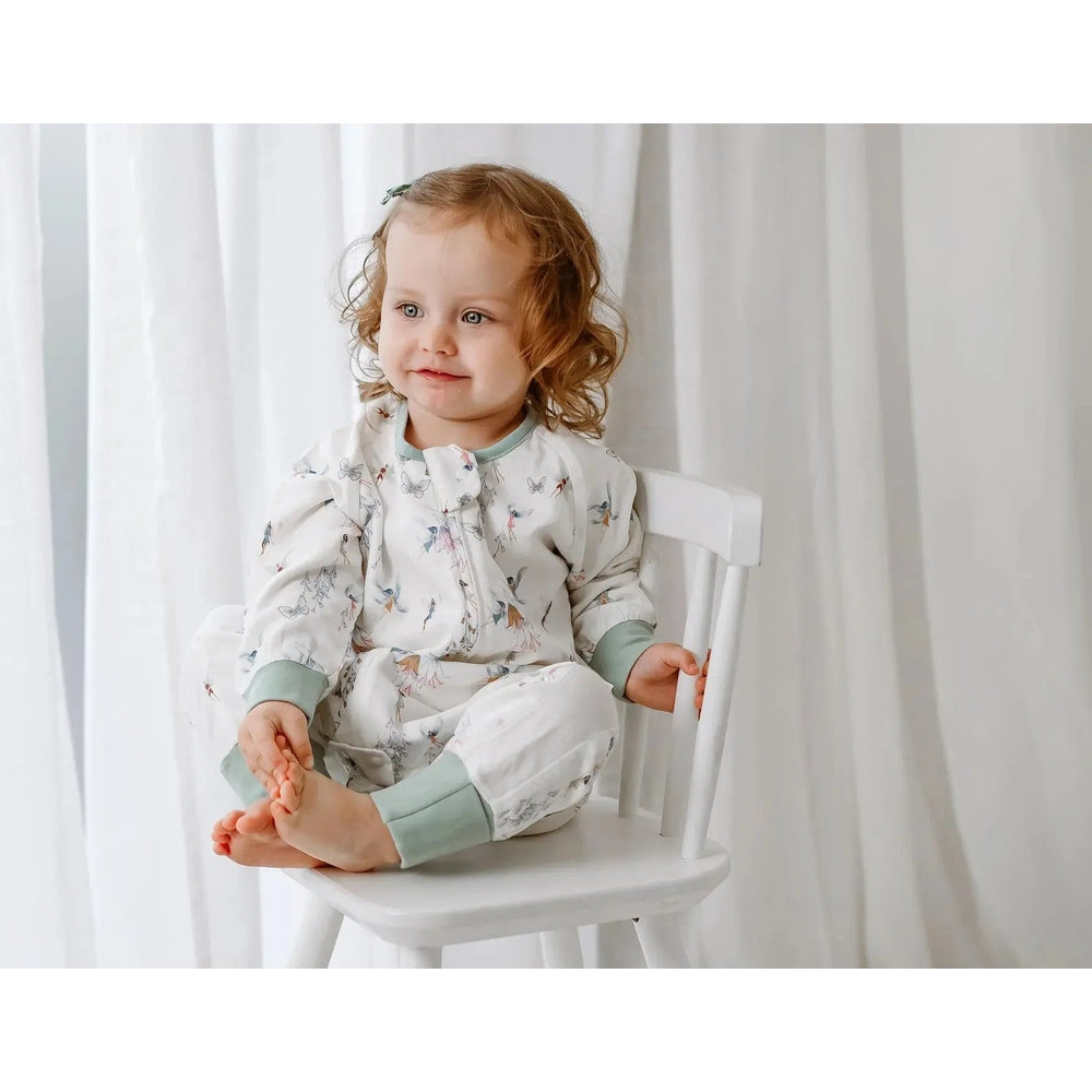 Nest Designs Bamboo Pima long-sleeve footed sleep bag, 0.6 Tog, fairy tale print; toddler sits on a white chair.