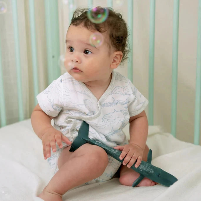 Nest Designs Bamboo Short Sleeve Kimono Onesie Surf's Up; baby sits in a crib, white with blue waves, holding a teal airplane.