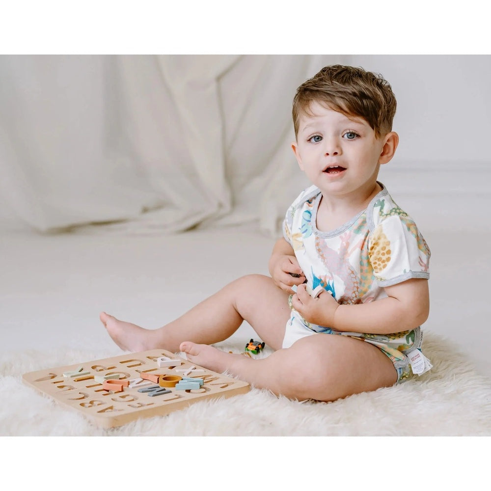 Nest Designs Bamboo Short Sleeve Onesie Dragon Dance worn by a baby sitting on a rug, pastel dragon print and soft fabric.