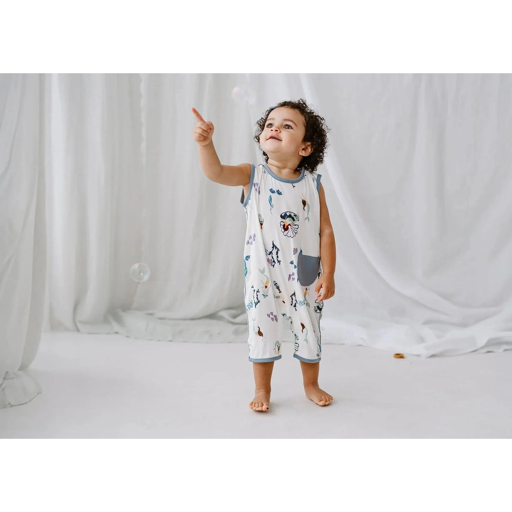 Nest Designs Bamboo Sleeveless Romper Splish Splash: toddler in white romper with blue trim and sea-creature print, blue pocket, pointing upward.