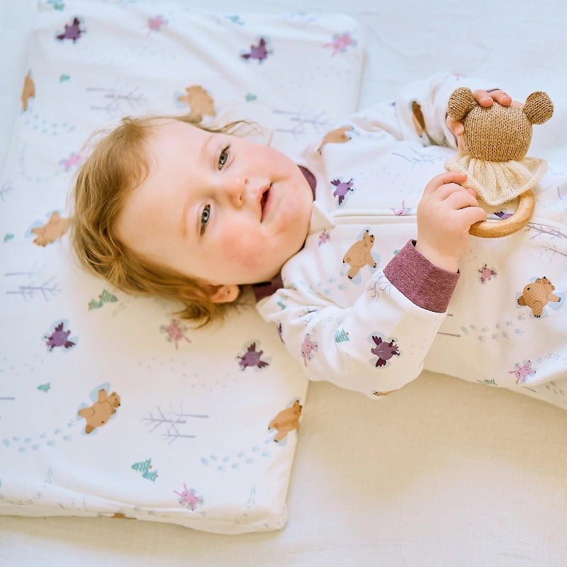 Nest Designs Bamboo Toddler Pillow with Pillowcase Snow Angels; baby smiles on a soft bamboo pillow while cuddling a small knitted bear.