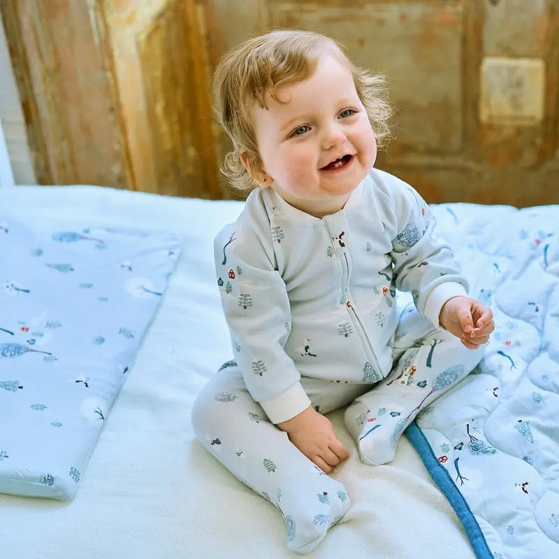 Nest Designs Bamboo Toddler Pillow With Pillowcase Snowy Snuggles; smiling baby sits on soft blue bedding, showing the cozy bamboo-filled pillow.