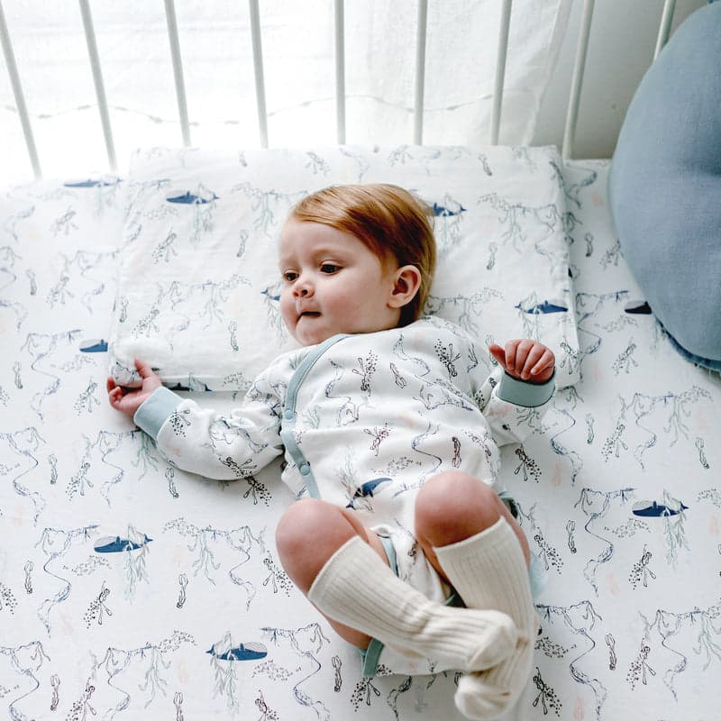 Nest Designs Bamboo Toddler Pillowcase Whale on a white bed, with a baby resting on whale-pattern bedding.