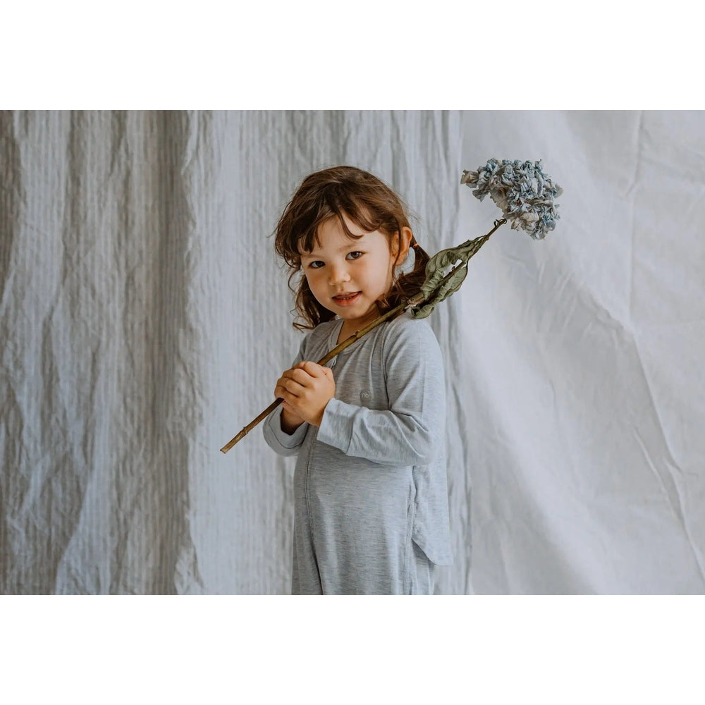 Nest Designs Basics bamboo cotton long-sleeve drop-bottom romper in grey dawn, worn by a toddler holding a dried flower.