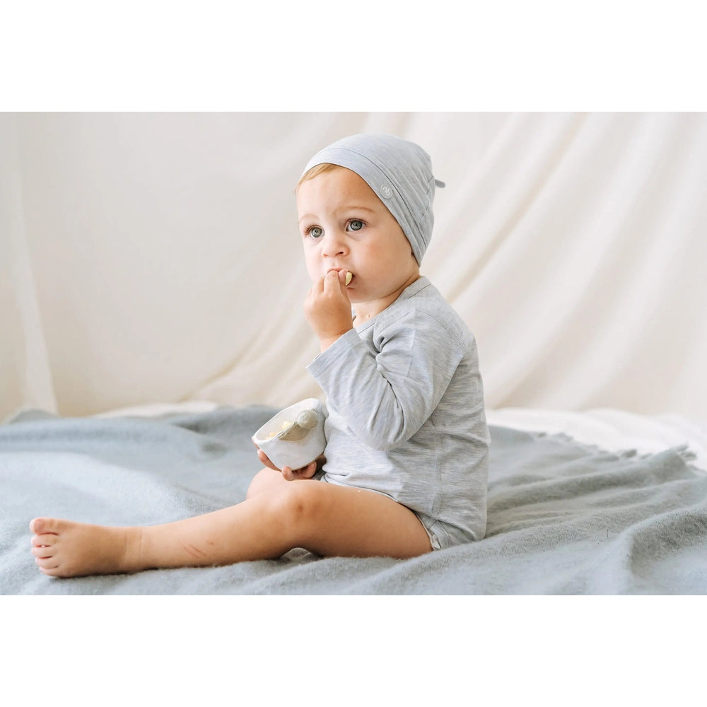 Nest Designs Basics Bamboo Cotton Long Sleeve Kimono Onesie Grey Dawn worn by baby sitting on a soft grey blanket, nibbling a snack.