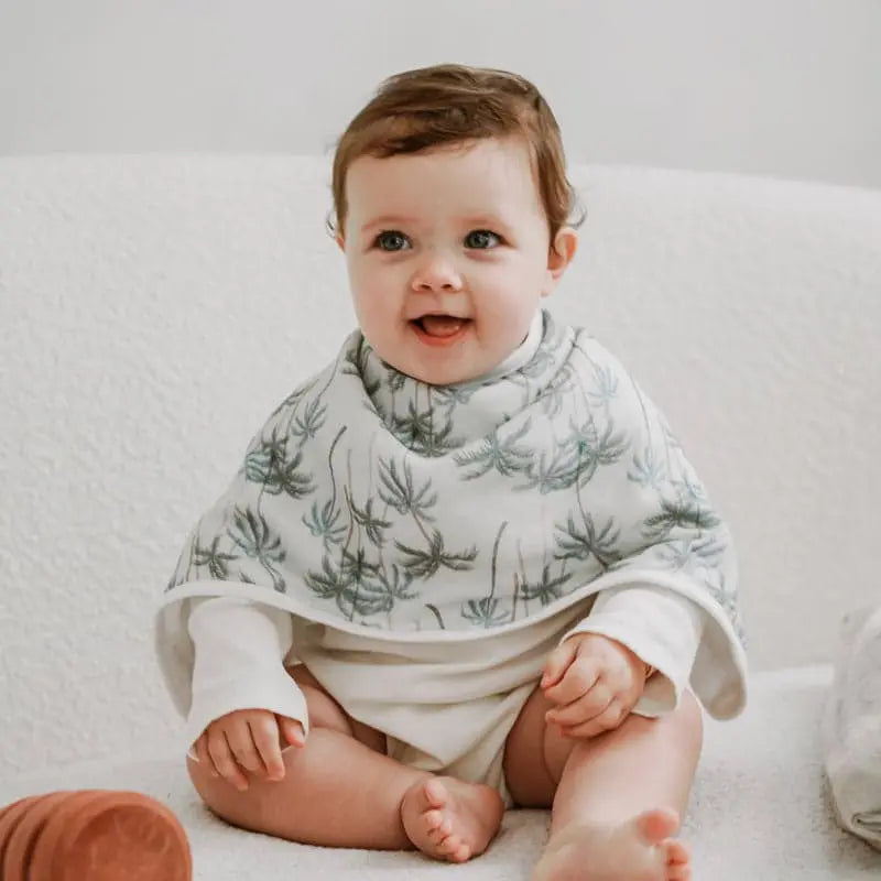 Nest Designs Burp'N Bib (2 Pack) baby wearing white bib with green palm print, seated on a white couch.