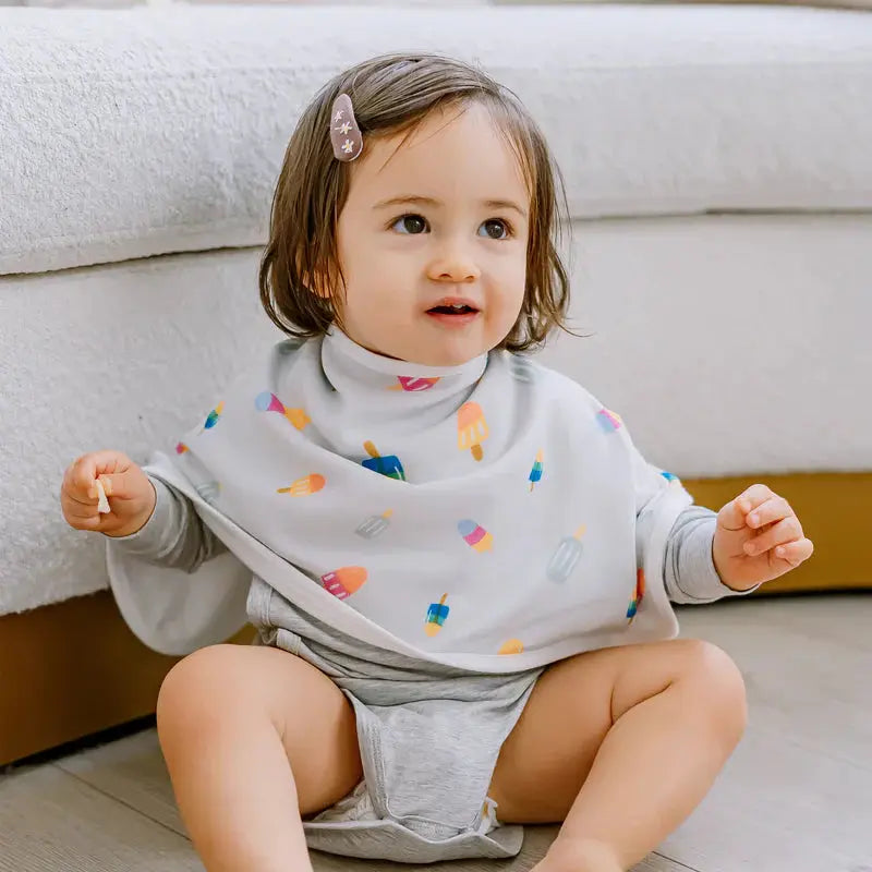 Nest Designs Burp'N Bib 2-pack; white bibs with colorful popsicles, worn by a smiling toddler sitting on the floor.