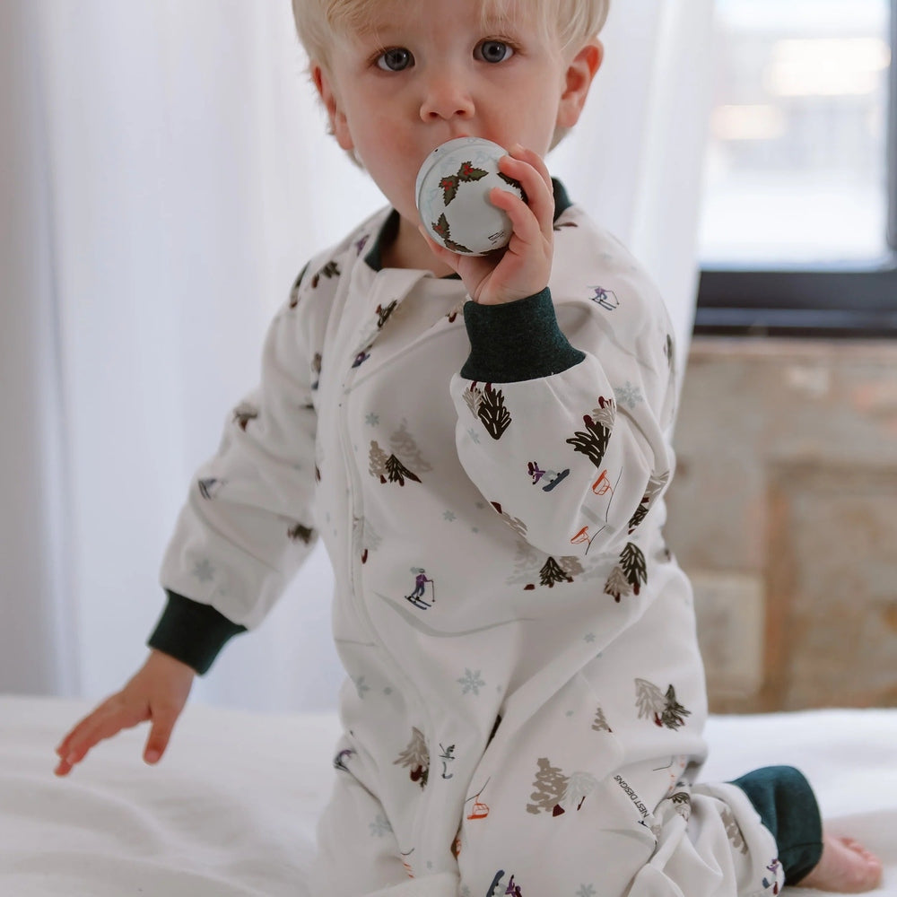 Nest Designs Cotton Long Sleeve Footed Sleep Bag 1.0 Tog, white with winter trees; toddler kneels holding a decorative ball.